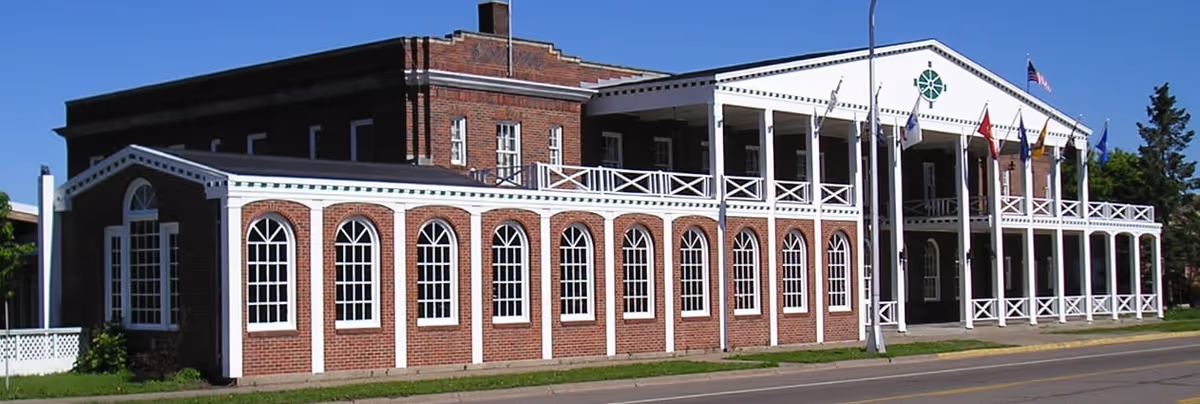 A large two-story brick building with white trim and multiple arched windows on the lower level. The building has a covered porch with white railings and several flags displayed on poles in front. The sky is clear and blue.