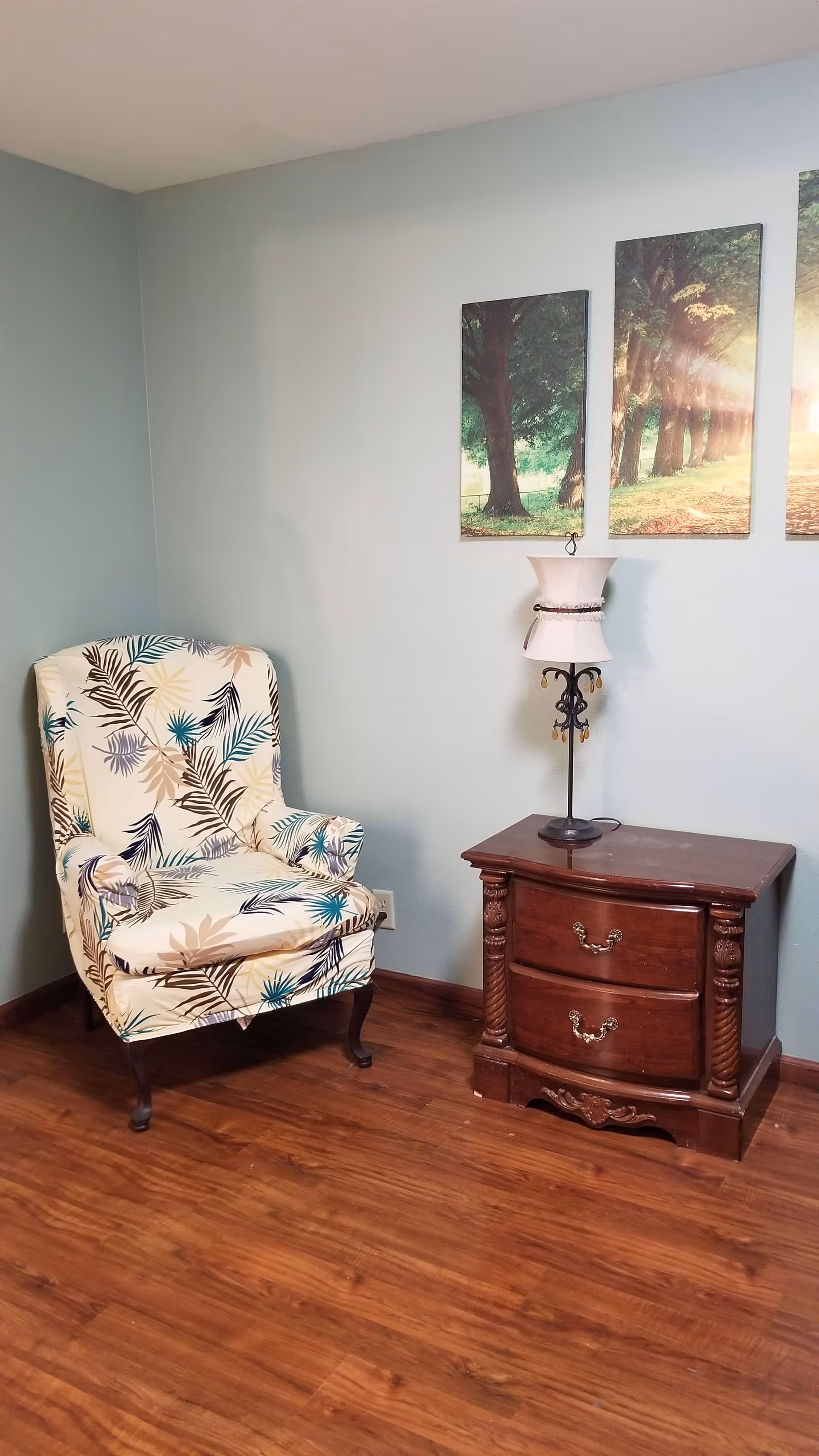 A corner of a room with a patterned armchair featuring leaf designs, a wooden side table with two drawers, and a decorative lamp on top. Above the table are three vertical wall art panels depicting a sunlit forest path with trees.