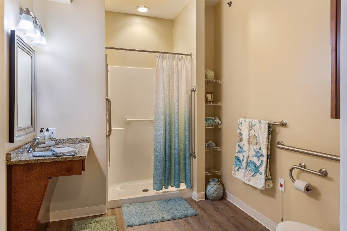 A bathroom with a walk-in shower featuring a blue and white gradient shower curtain, grab bars, and a built-in seat. There is a wooden vanity with a granite countertop, a sink, and a mirror with lights above it. The bathroom has beige walls, wood-look flooring, a towel rack with a sea-themed towel, and open shelving with decorative items.