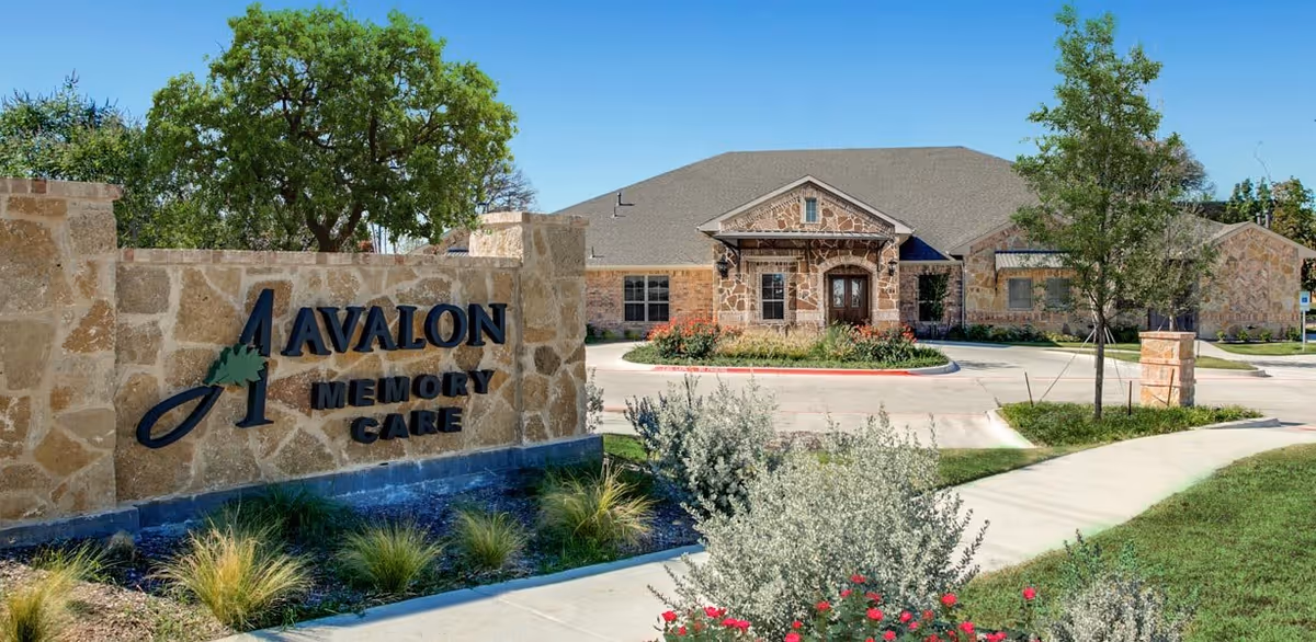 Exterior view of Avalon Memory Care facility showing a stone sign with the facility name and a large building with stone facade, surrounded by trees, shrubs, and a clear blue sky.