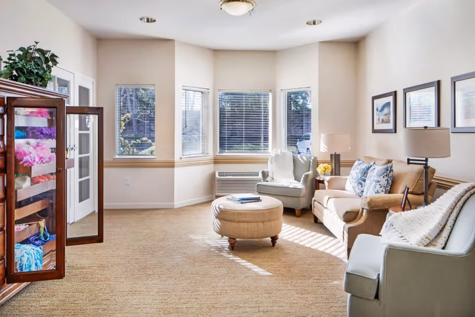 A bright and cozy living room with beige walls and carpet. The room features a beige sofa with patterned cushions, two armchairs with blankets draped over them, a round ottoman in the center, and a wooden cabinet with glass doors displaying colorful folded blankets. Three framed pictures hang on the wall above the sofa, and large windows allow natural light to fill the space.