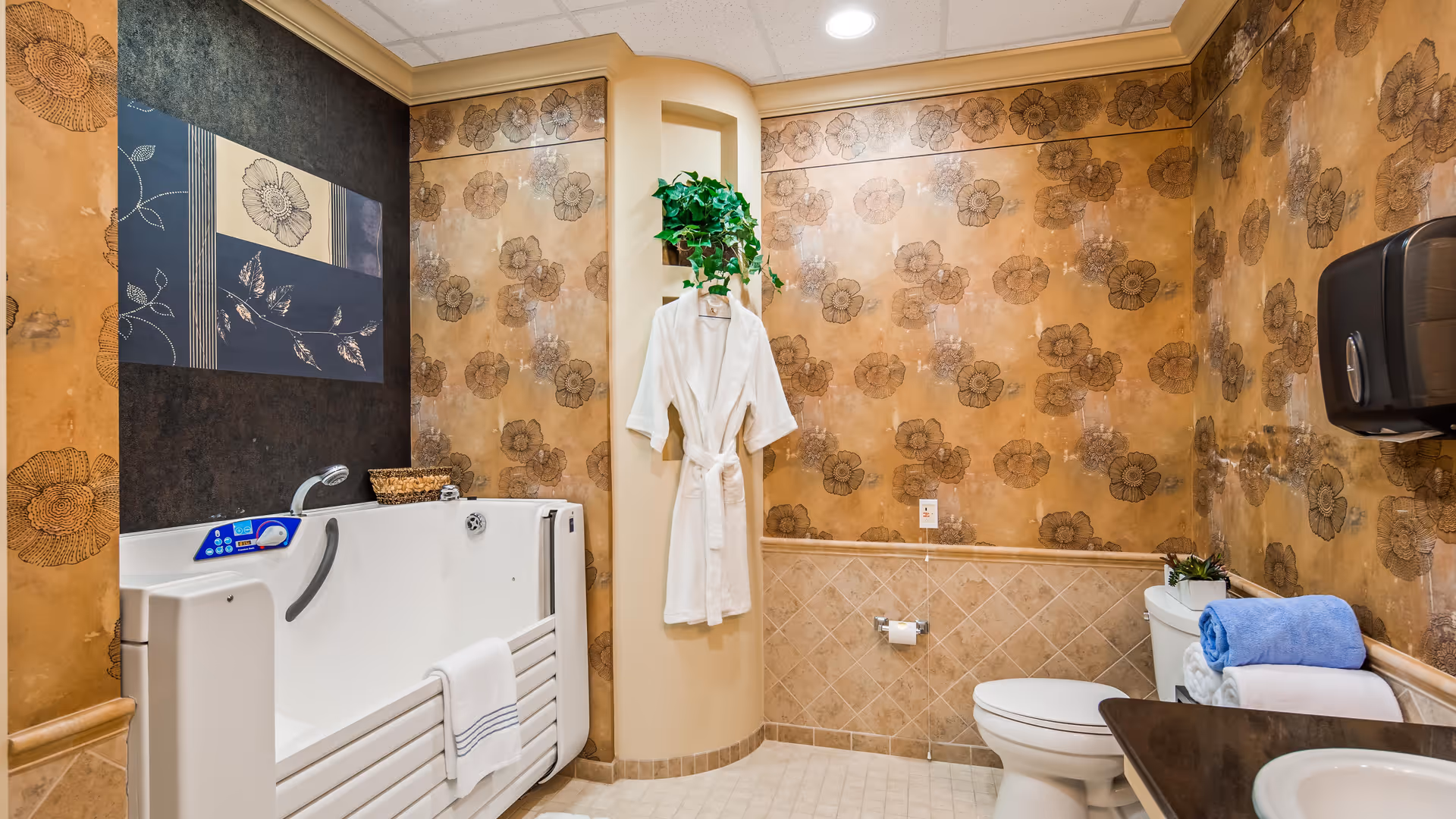 A bathroom with floral patterned wallpaper and beige tiled lower walls. The room features a white walk-in bathtub with a towel hanging on its side, a white toilet with folded towels and a small plant on top, a white bathrobe hanging on a wall hook, a black paper towel dispenser, and a dark countertop with a white sink. There is also a decorative framed artwork on the wall above the bathtub.