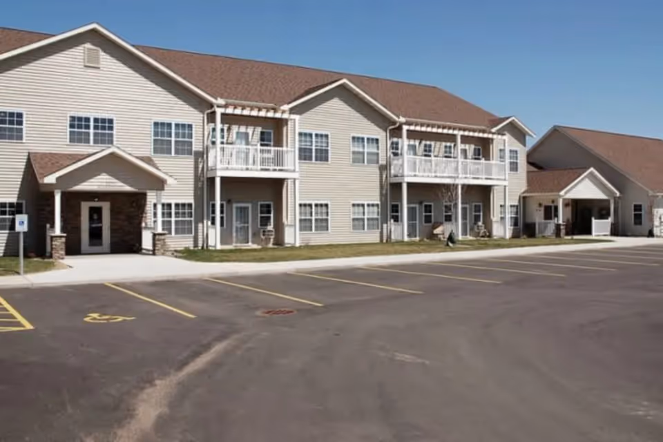 Exterior view of a two-story senior living facility building with beige siding and brown roofs, featuring balconies and a parking lot with marked spaces including a handicapped spot.