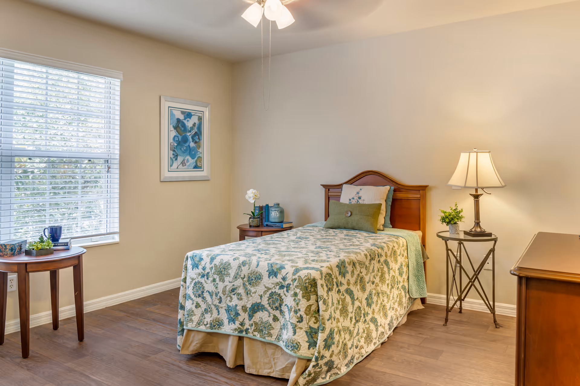 A cozy bedroom with a single bed covered in a floral patterned quilt in shades of green and blue. The bed has multiple pillows and a wooden headboard. To the right of the bed is a small round metal side table with a lamp and a small plant. To the left of the bed is a wooden nightstand with decorative items including books and a small plant. A window with white blinds lets in natural light, and a framed artwork with blue flowers hangs on the wall. The floor is wooden, and the walls are painted a light beige color.