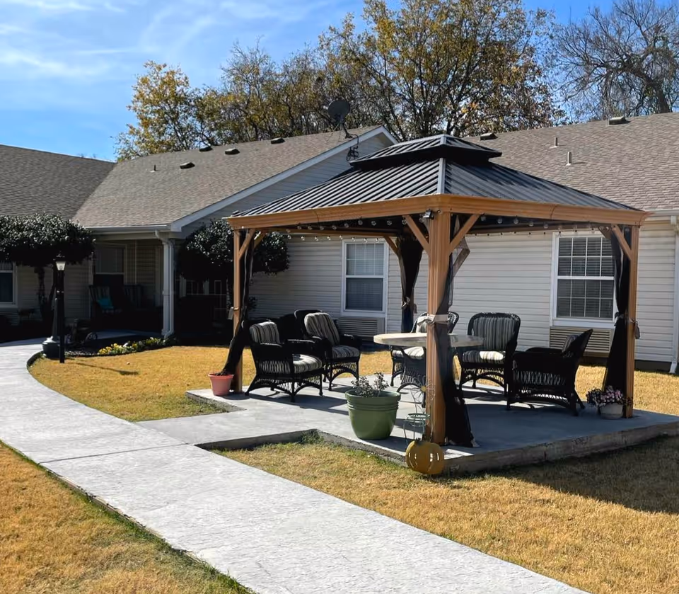 Outdoor seating area with a wooden gazebo featuring a metal roof, surrounded by cushioned wicker chairs and a round table. The gazebo is situated on a concrete slab with potted plants around it, adjacent to a building with beige siding and multiple windows. A curved concrete walkway leads to the gazebo, and trees with autumn foliage are visible in the background under a clear blue sky.