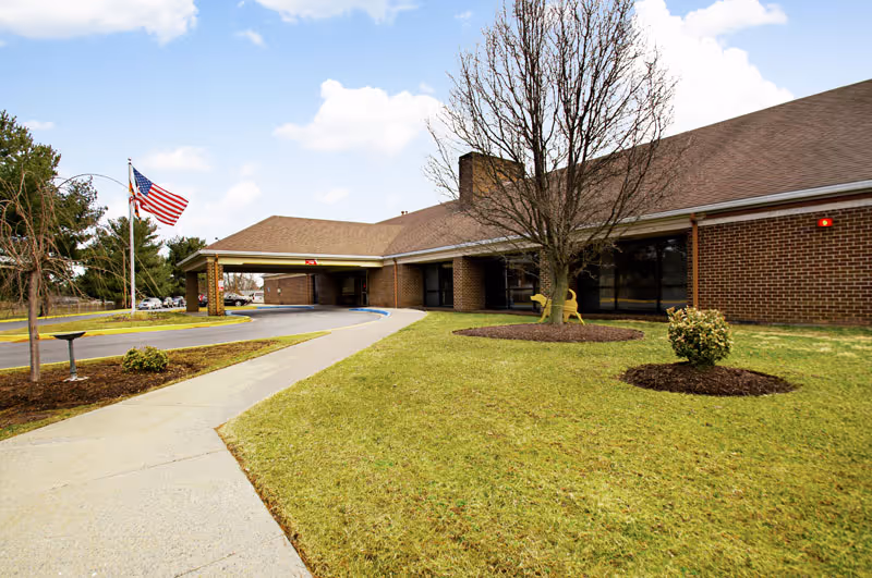 Front entrance of a single-story brick senior living building with a covered drive, walkway, flagpole, and lawn.