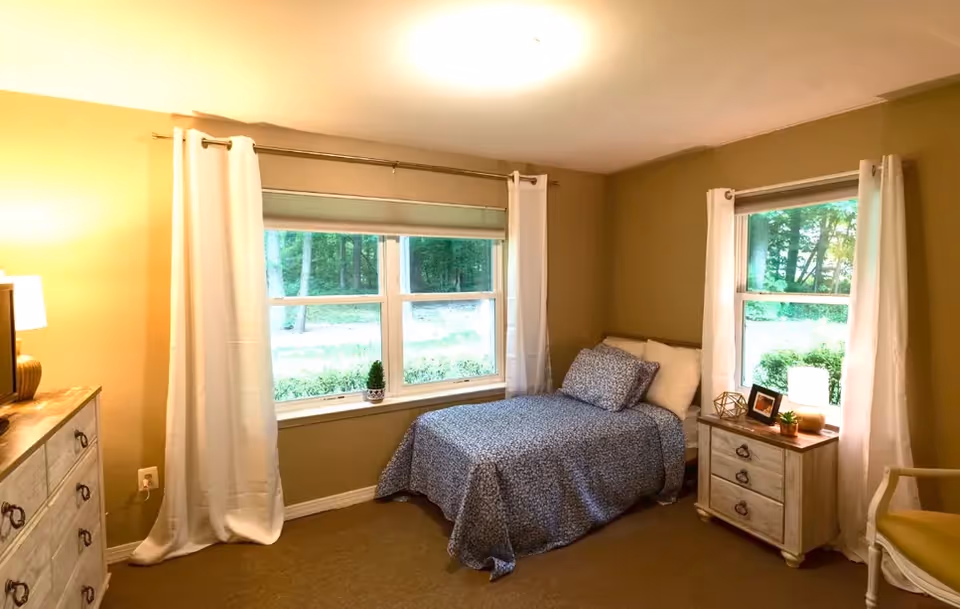 A cozy bedroom with a single bed covered in a blue patterned bedspread and matching pillow. The room has two windows with white curtains, letting in natural light and showing a view of greenery outside. There is a wooden dresser with a lamp on the left side and a small wooden nightstand with a lamp, picture frame, and decorative items on the right side. A chair with a yellow cushion is partially visible on the right.