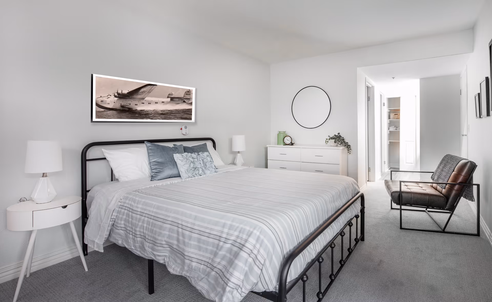 A bright, modern bedroom with a large bed featuring a black metal frame and striped bedding. There are two white bedside tables with lamps on either side of the bed. Above the bed hangs a framed black and white photo of an airplane. Across from the bed is a white dresser with a round mirror above it, a clock, and a small plant. To the right, there is a black metal-framed chair with a cushion and a throw blanket. The room has light gray carpeting and white walls, with a hallway leading to another room in the background.