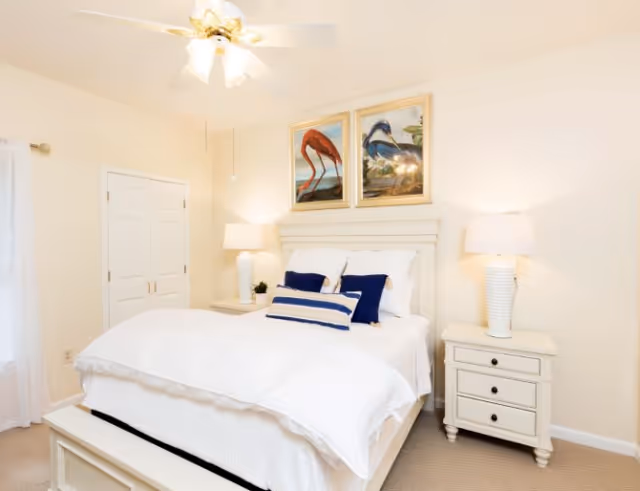 Bright white bedroom with a bed dressed in white linens, navy accent pillows, two bedside tables with lamps, a ceiling fan, and framed bird artwork above the headboard.