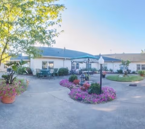 Outdoor courtyard area of a senior living facility with a paved walkway, flower beds filled with colorful flowers, green shrubs, patio tables and chairs, and a large umbrella. The building surrounds the courtyard with windows facing the outdoor space. A tree provides some shade on the left side.