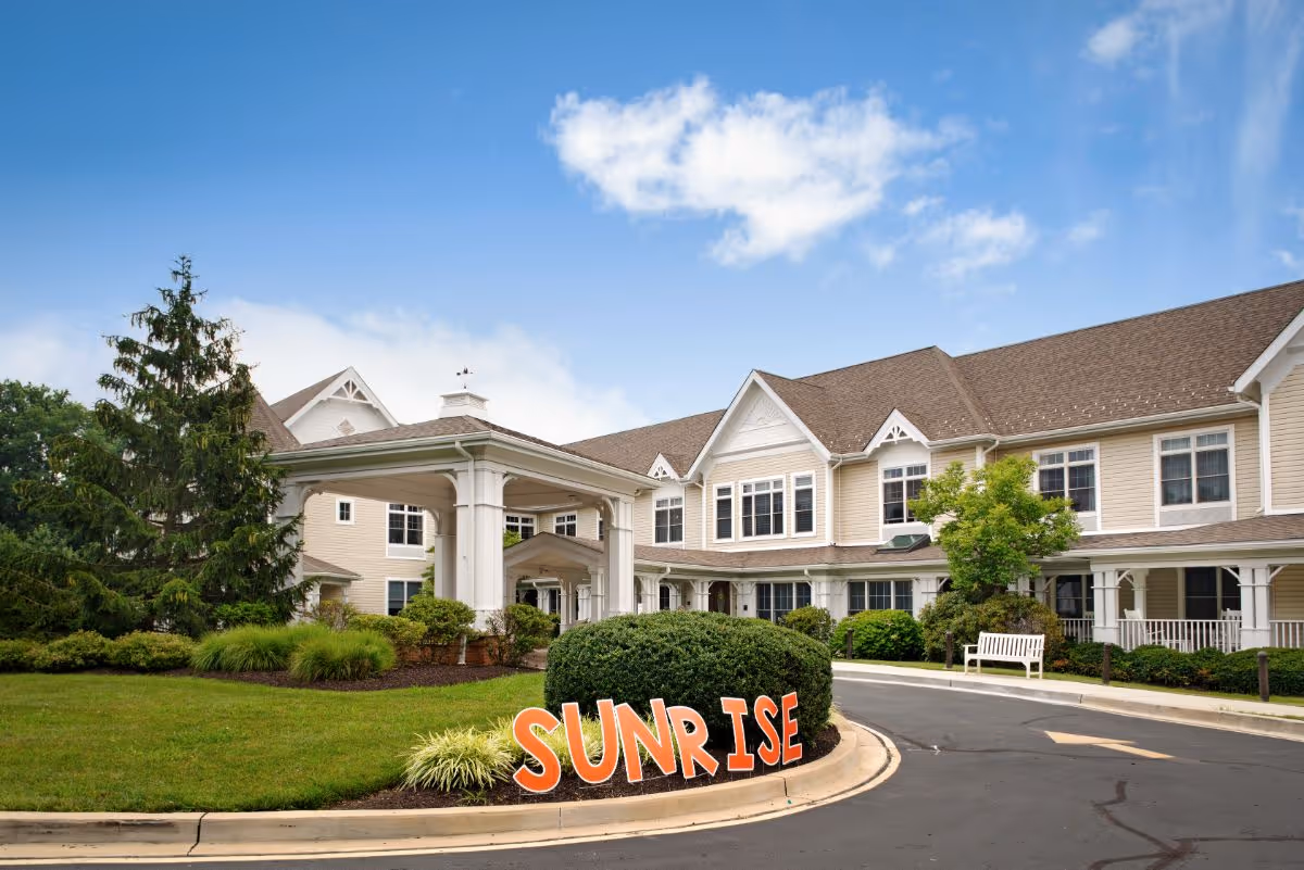 Exterior view of a senior living facility building with beige siding and white trim under a blue sky with some clouds. There is a covered entrance with columns, well-maintained landscaping including bushes and trees, and a curved driveway. Large orange letters spelling 'SUNRISE' are placed on the grass near the driveway.