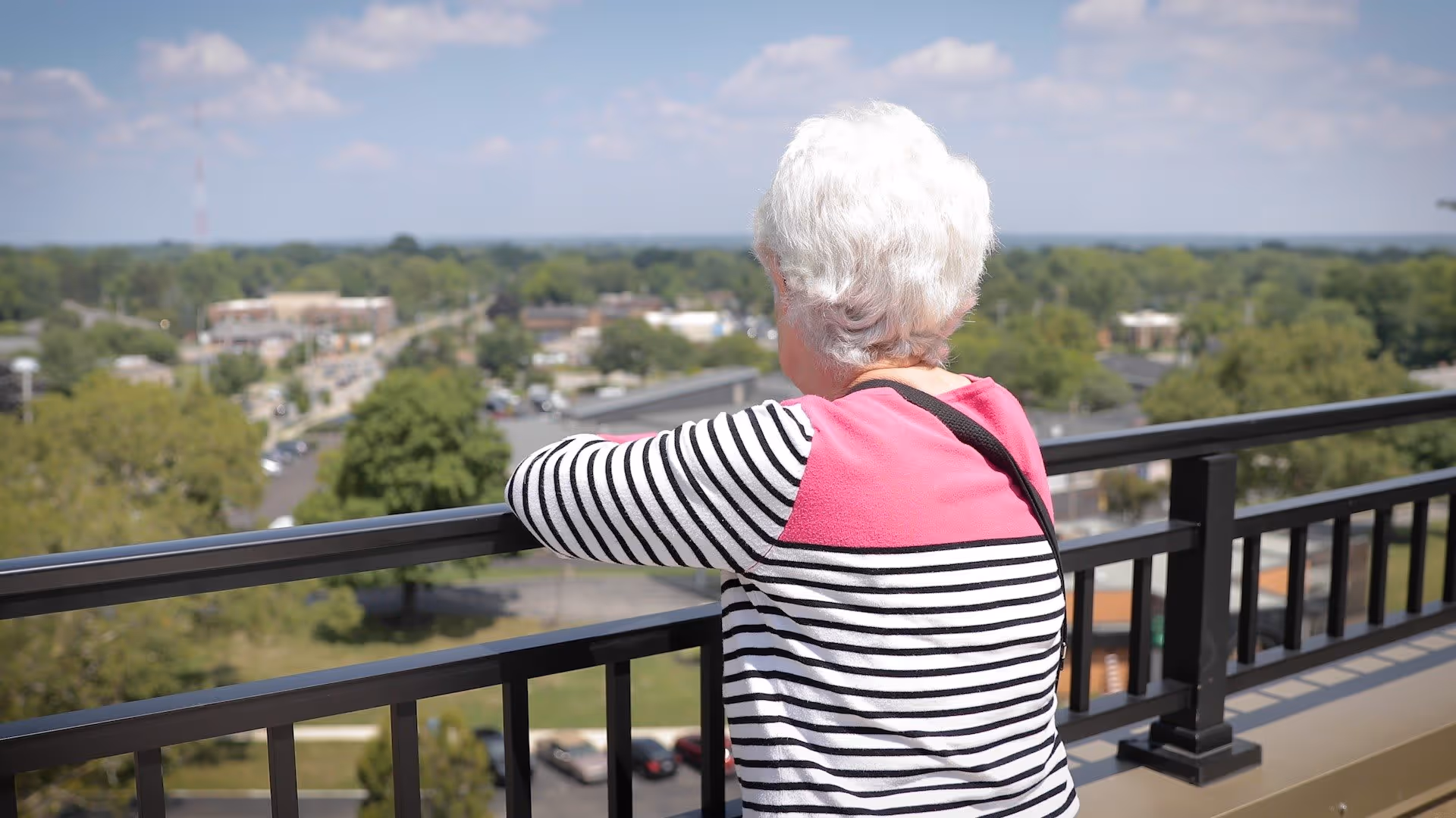 An elderly woman with white hair wearing a striped black and white shirt with a pink shoulder panel is standing on a balcony, looking out over a suburban area with trees, buildings, and a partly cloudy sky.