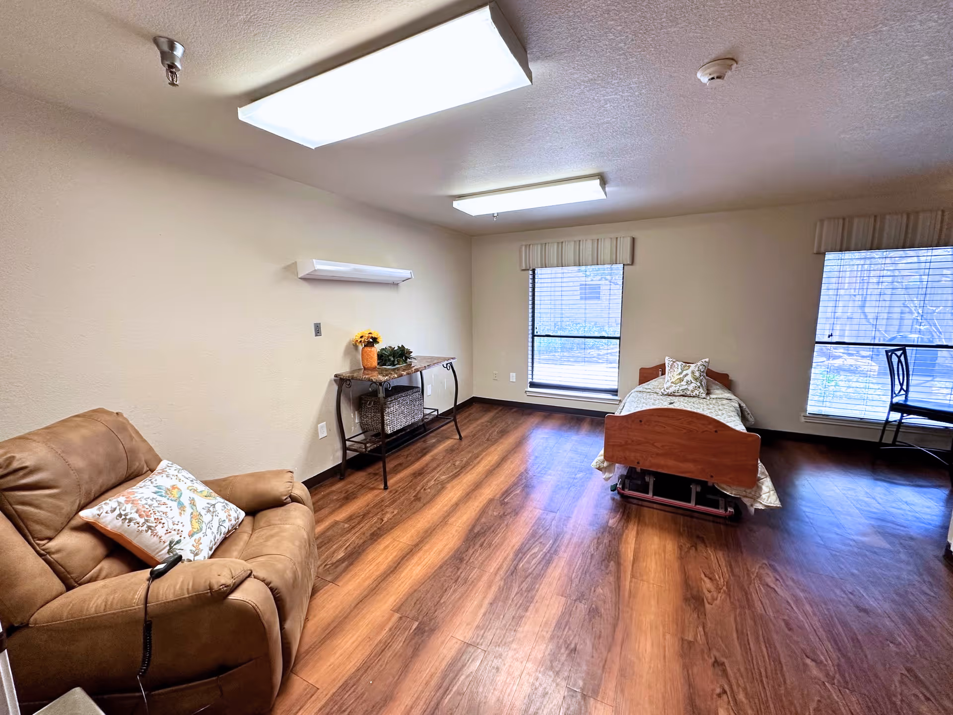 Spacious senior bedroom with a recliner, single bed, wood floors, windows, and a small side table.
