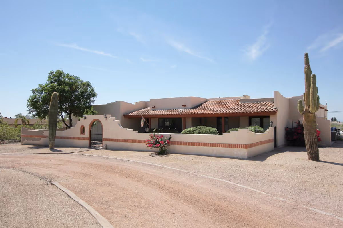 Exterior view of a single-story building with southwestern architectural style, featuring a tiled roof, stucco walls, a gated courtyard, desert landscaping with cacti and a tree, and a clear blue sky.
