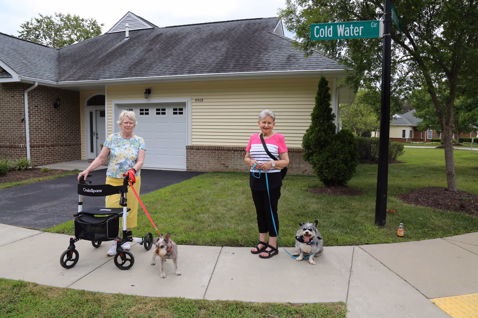 Two elderly women standing on a sidewalk in front of a house. One woman is using a walker and holding a small dog on a red leash. The other woman is holding a dog on a blue leash. There is a street sign that reads 'Cold Water Cir' and green grass and trees around them.