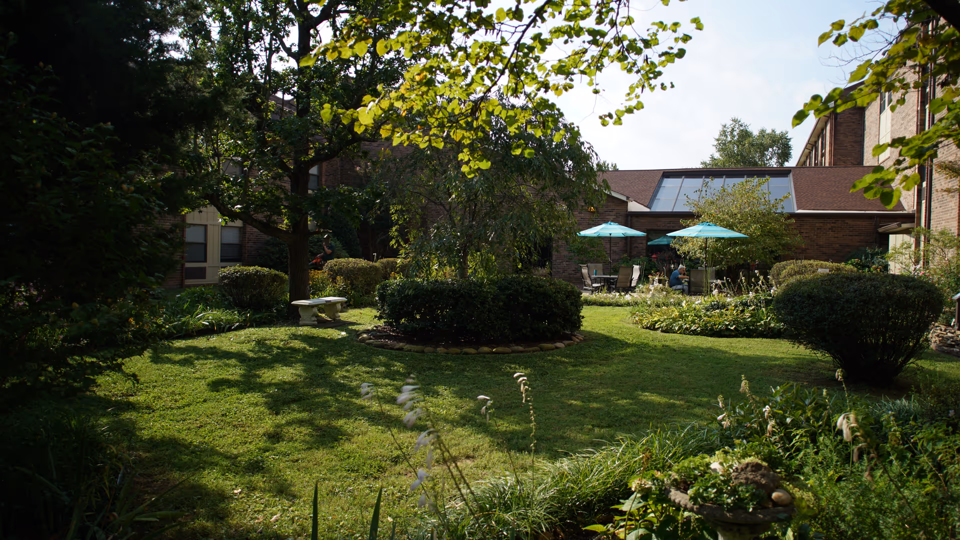 Courtyard garden with a grassy lawn, trees, shrubs, and a patio area with teal umbrellas beside a brick building.