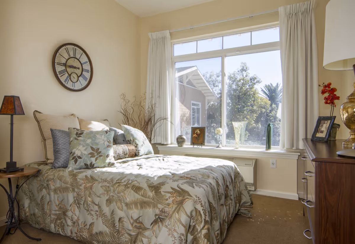 Bright bedroom with a patterned bedspread and pillows, a large window with decorative vases on the sill, and a wall clock and lamps.