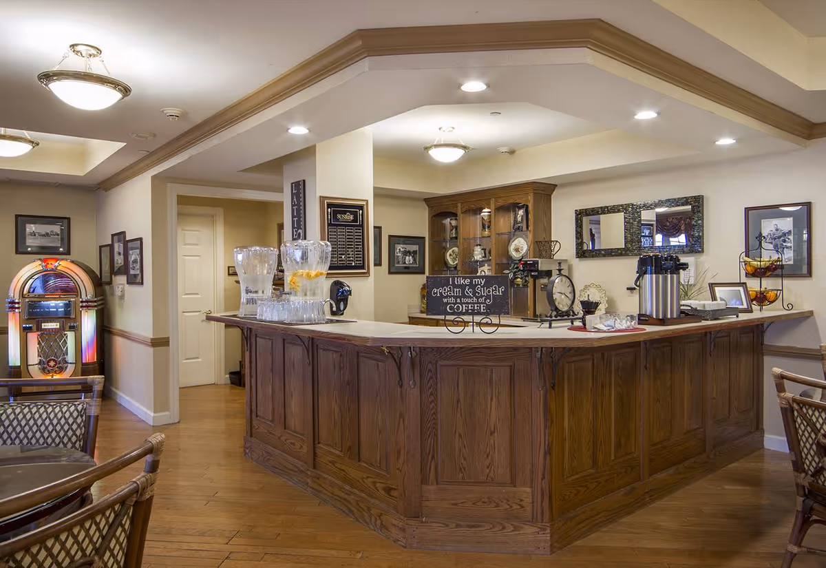 Interior view of a senior living facility's common area with a wooden counter featuring water pitchers, coffee dispensers, and decorative items. There is a jukebox against the wall, framed pictures, and a wooden cabinet with plates and trophies. The room has warm lighting and wooden flooring.