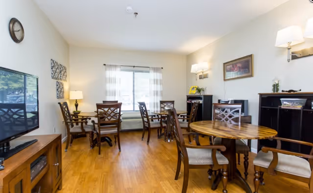A well-lit common area with wooden floors featuring multiple round wooden tables and chairs arranged for seating. There is a large window with sheer curtains letting in natural light, a wall clock, a flat-screen TV on a wooden stand, wall-mounted lamps, and framed artwork on the walls.