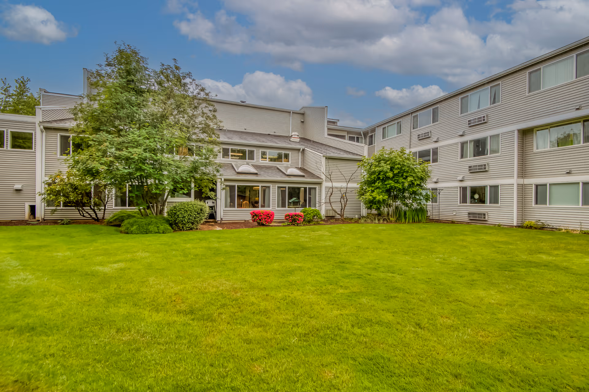 Green lawn and landscaped shrubs in a courtyard in front of a light-colored multi-story apartment building.