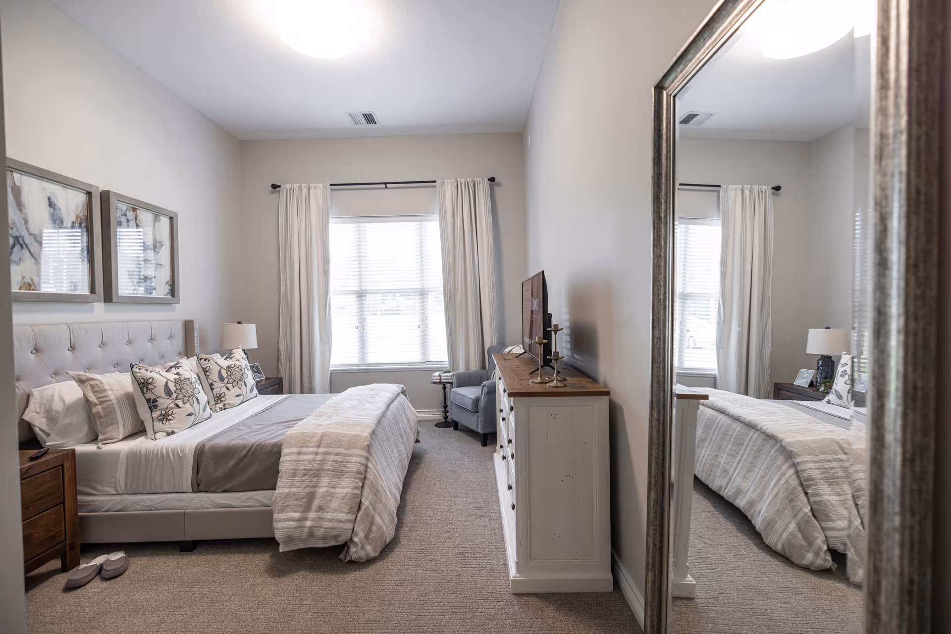 Neutral-toned bedroom with a tufted headboard bed, dresser, armchair, window with curtains, and a large wall mirror.