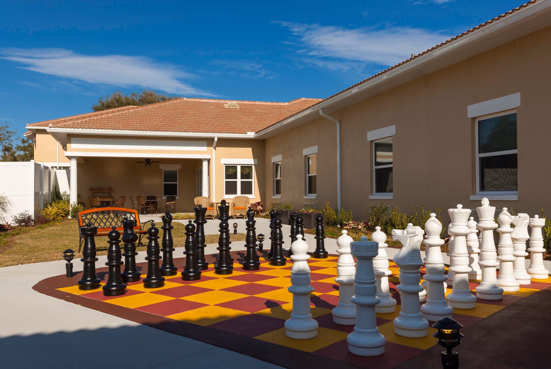 Outdoor area of a senior living facility featuring a large chessboard with oversized black and white chess pieces set up for a game. The area is paved with concrete and surrounded by a beige building with several windows. There is a covered patio with chairs and a bench nearby, under a clear blue sky.