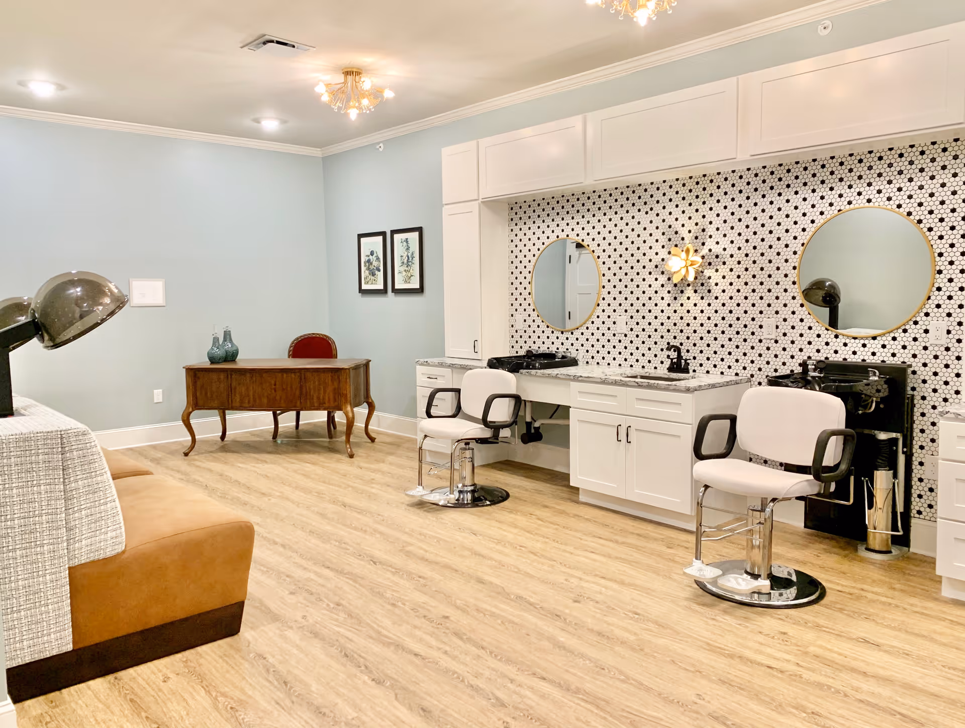 Interior room with two white salon chairs in front of a counter with sinks and round mirrors mounted on a wall with black and white patterned tiles. There is a wooden desk with a red chair and decorative vases against a light blue wall. The floor is light wood, and the ceiling has a small chandelier and recessed lighting.