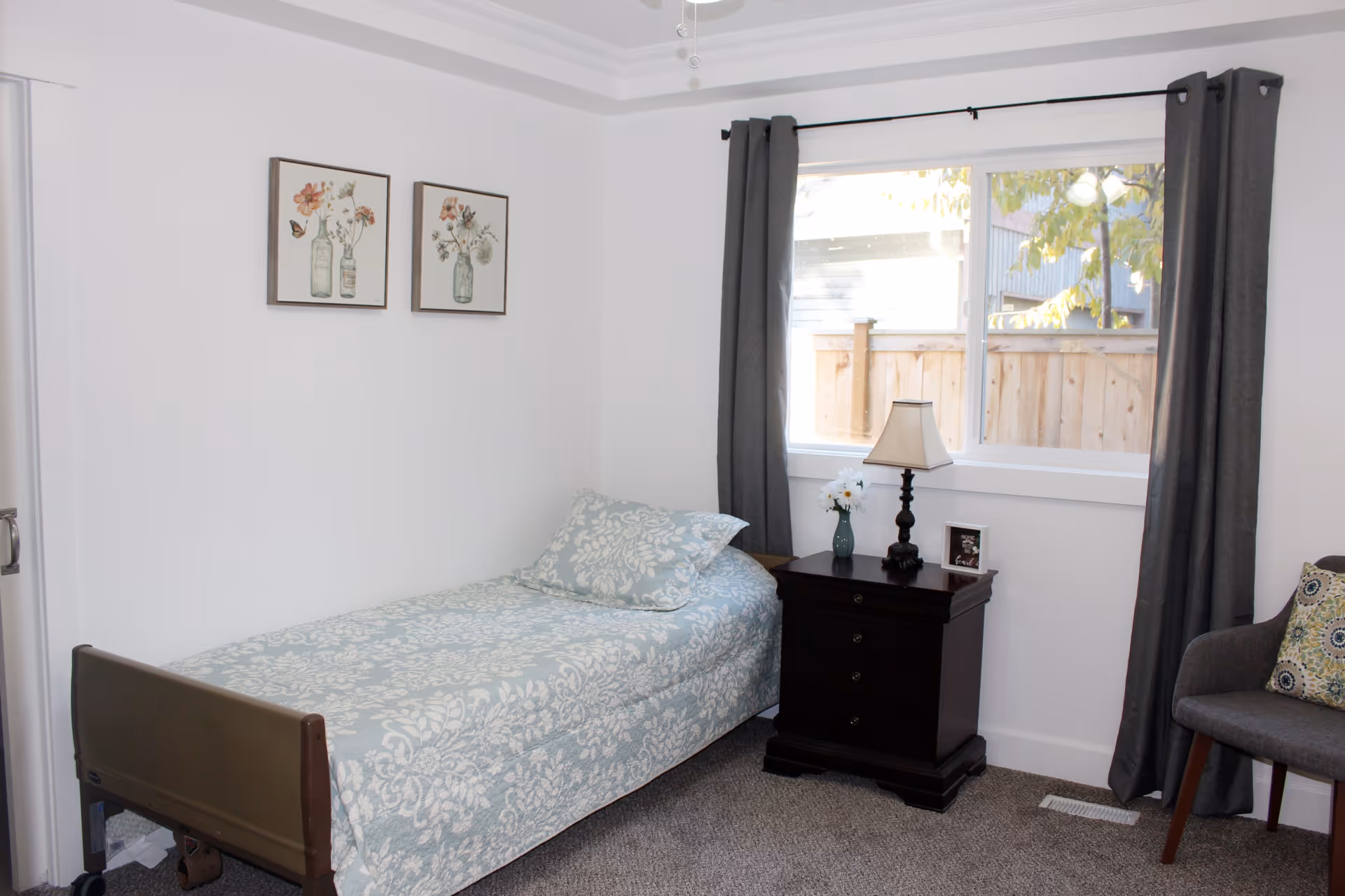 A small, neatly arranged bedroom with a single bed covered in a light blue patterned bedspread and matching pillow. Next to the bed is a dark wooden nightstand with a lamp, a small vase with flowers, and a framed photo. A window with gray curtains lets in natural light, and two framed floral prints hang on the white wall above the bed. A gray upholstered chair with a decorative pillow is positioned near the window.