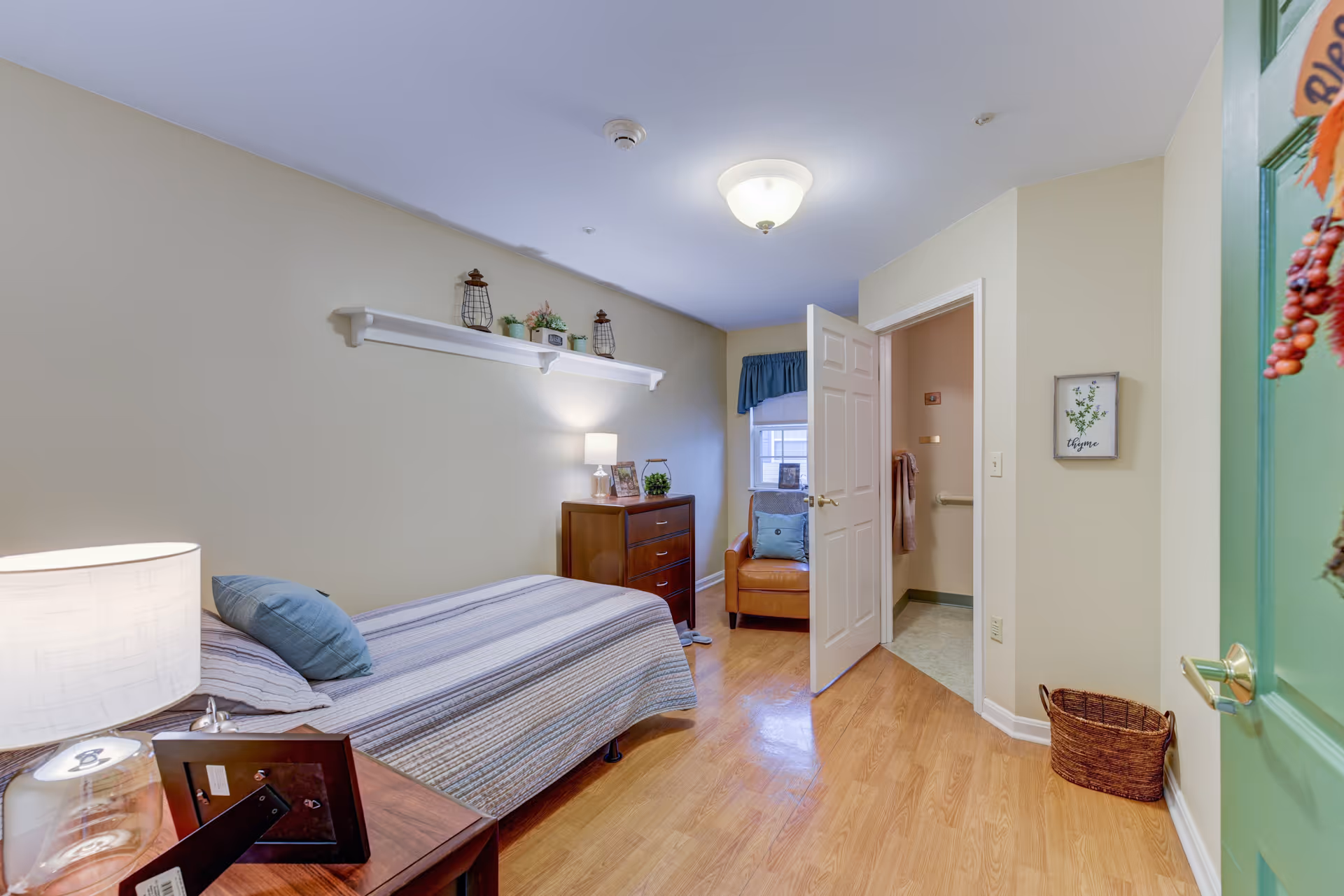 A small, cozy bedroom with a single bed covered in a striped quilt and a blue pillow. Next to the bed is a wooden nightstand with a lamp and a picture frame. Across from the bed is a wooden dresser with a lamp, framed photos, and small plants on top. A leather armchair with a blue pillow is positioned near a window with a blue valance. The door to an adjacent bathroom is open, showing a towel hanging on a grab bar. The room has light beige walls, wood flooring, and a green door partially visible on the right side with a decorative hanging.