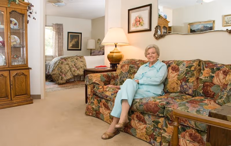 An elderly woman in light blue pajamas sitting and smiling on a floral patterned couch in a cozy living room area. Behind her, there is a wooden cabinet with decorative plates, a lamp on a side table, and a bedroom with a bed covered in a floral bedspread visible through an open doorway.