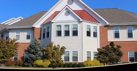 Senior living facility exterior with beige siding, brick accents, and landscaped trees.