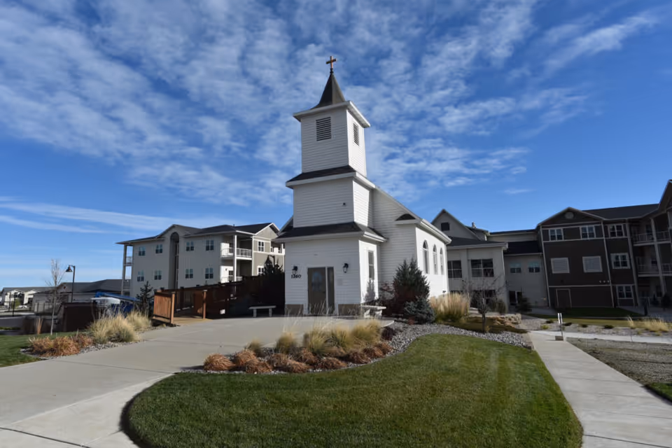 Exterior view of a white chapel with a steeple and cross, surrounded by landscaped grass and shrubs, with multi-story residential buildings in the background under a partly cloudy blue sky.