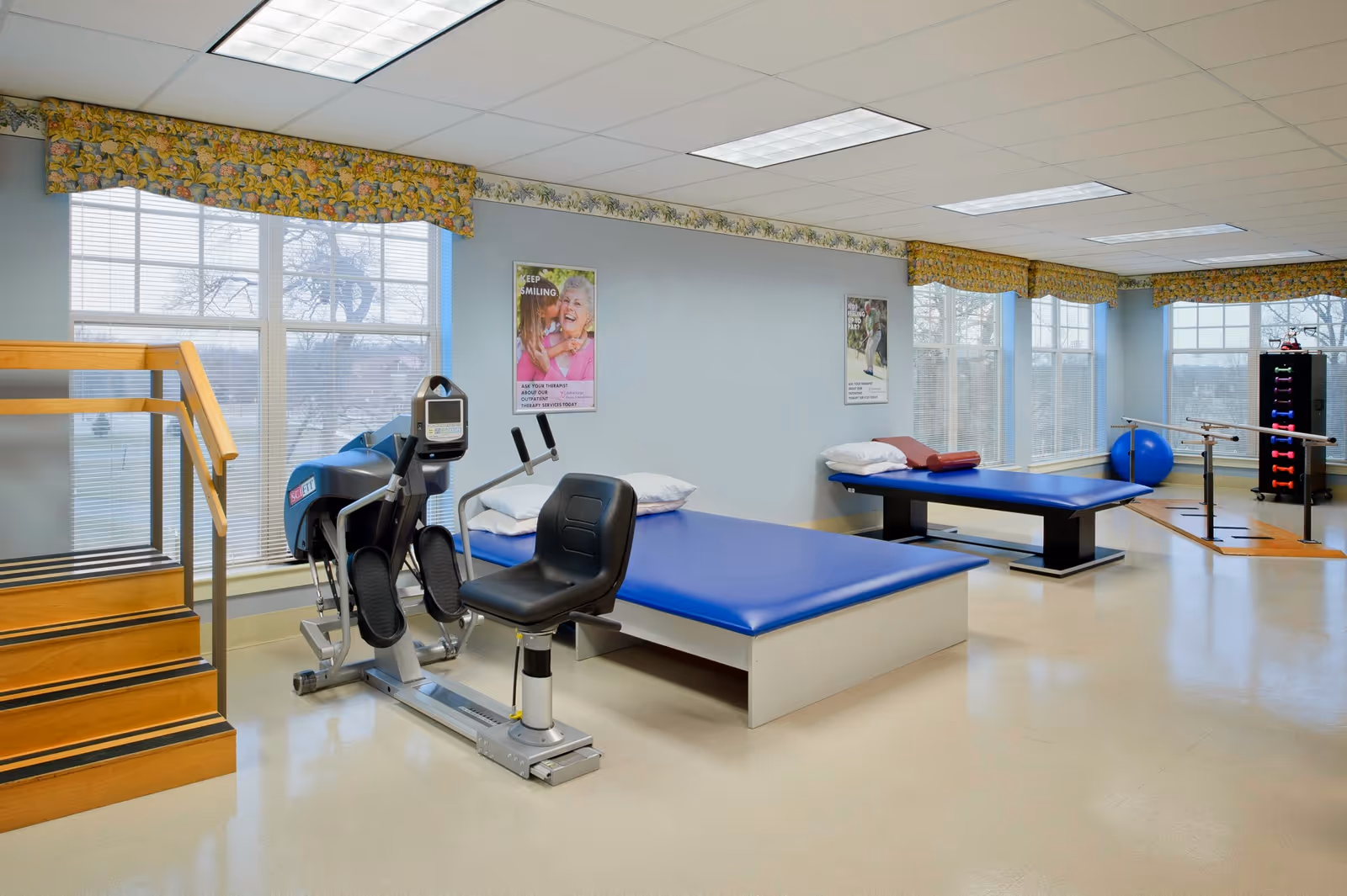A physical therapy room with exercise equipment including a recumbent bike, two padded therapy tables with pillows, parallel bars for walking exercises, a blue exercise ball, and a rack of colorful dumbbells. The room has large windows with floral valances, light blue walls, and a staircase with wooden steps and handrails.