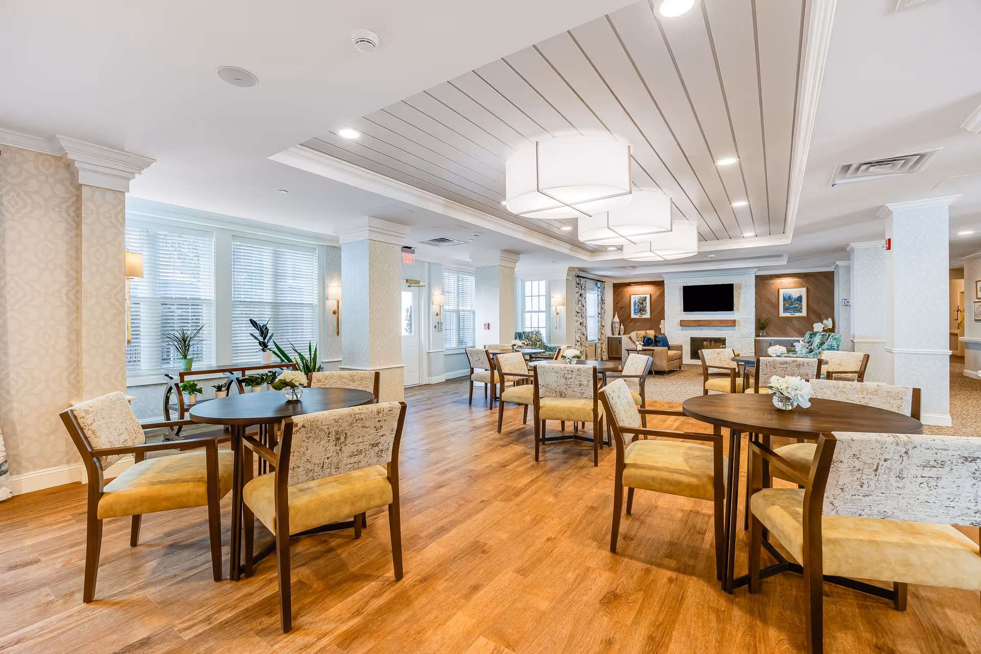 Bright, spacious communal dining and lounge area with round tables and upholstered chairs, hardwood floors, and a fireplace with a TV in the background.