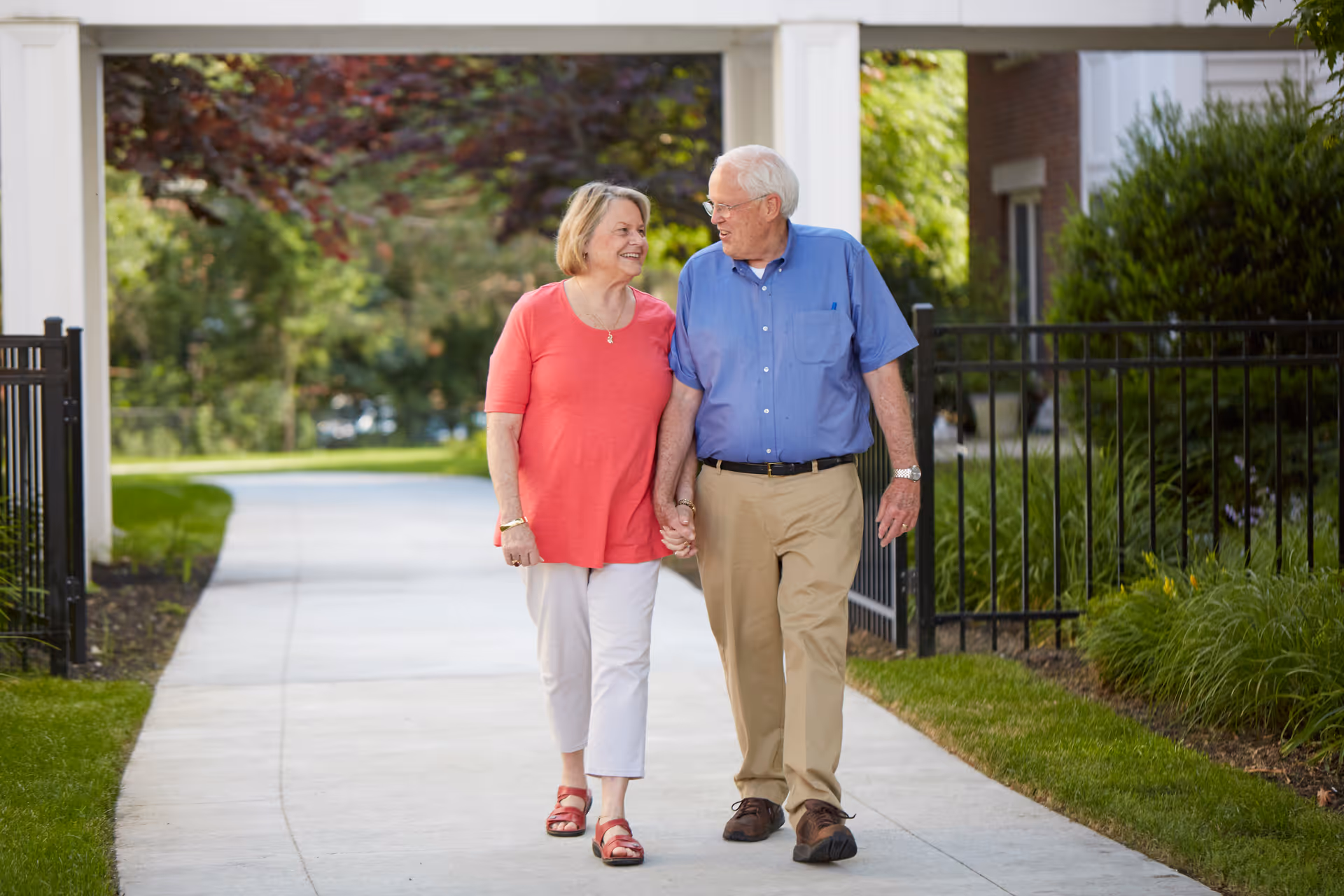 An elderly couple holding hands and walking along a paved pathway outdoors, surrounded by greenery and a black metal fence. The woman is wearing a coral top and white pants, and the man is wearing a blue shirt and beige pants. They are smiling and looking at each other.