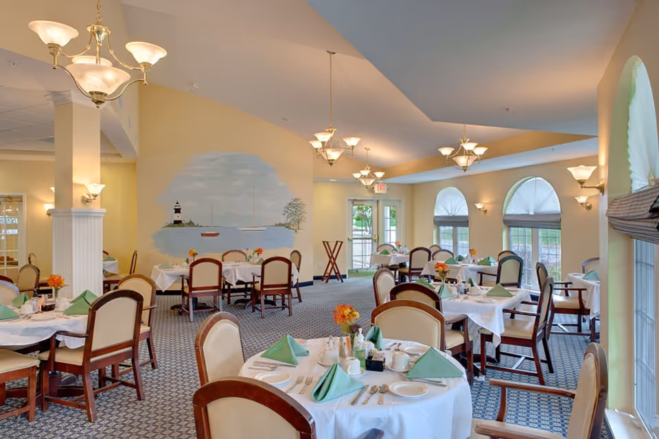 A bright dining room with multiple round tables covered with white tablecloths, set with plates, cups, silverware, and green folded napkins. The room has beige walls, carpeted floors, and several chandeliers hanging from the ceiling. Large windows with arched tops and pleated shades let in natural light. A mural of a lighthouse and sailboats on the water is painted on one wall.
