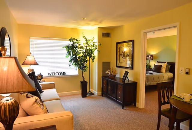 A warmly lit living room with a beige sofa adorned with patterned pillows, a table lamp, a potted plant near a window with blinds, and a decorative 'WELCOME' sign on the windowsill. A dark wooden sideboard with decorative items and a framed picture is against the wall. Through an open doorway, a bedroom with a bed, pillows, and a bedside lamp is visible. A small dining table with chairs is partially visible on the right.
