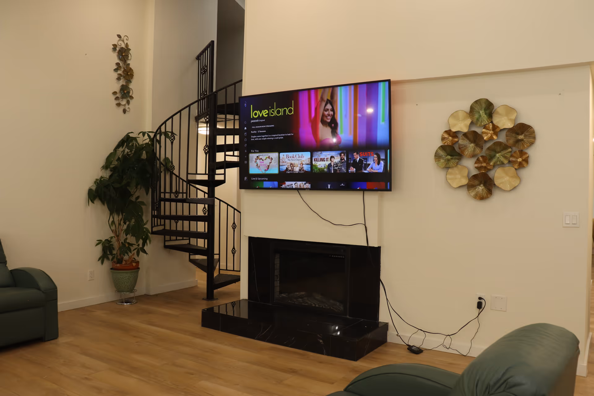 A living room with a mounted flat-screen TV displaying the show Love Island. Below the TV is a black fireplace with a marble base. To the left of the fireplace is a black spiral staircase and a large potted plant. The room has light-colored walls and wooden flooring. There is decorative wall art on both sides of the fireplace and green upholstered furniture partially visible on the left and right edges of the image.