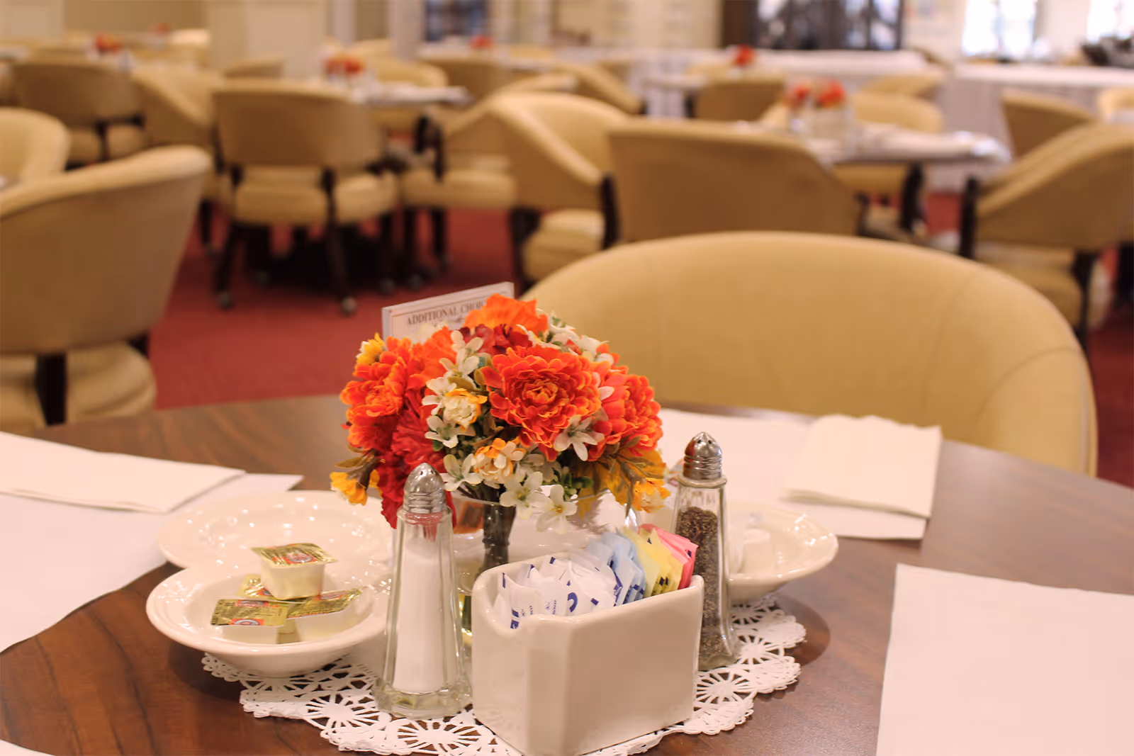 A round wooden dining table set with white plates, napkins, salt and pepper shakers, a container with sugar packets, and a small vase with red, orange, and white flowers. In the background, there are multiple similar tables and beige upholstered chairs on a red carpeted floor, suggesting a communal dining area.