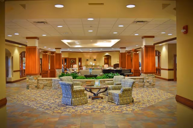Spacious and well-lit common area in a senior living facility with patterned carpet and tiled floor. The room features a circular seating arrangement with four upholstered armchairs around a small wooden table. In the background, there are wooden columns with stone bases, green plants, and additional seating areas including sofas and a piano. The ceiling has recessed lighting and a decorative central panel.
