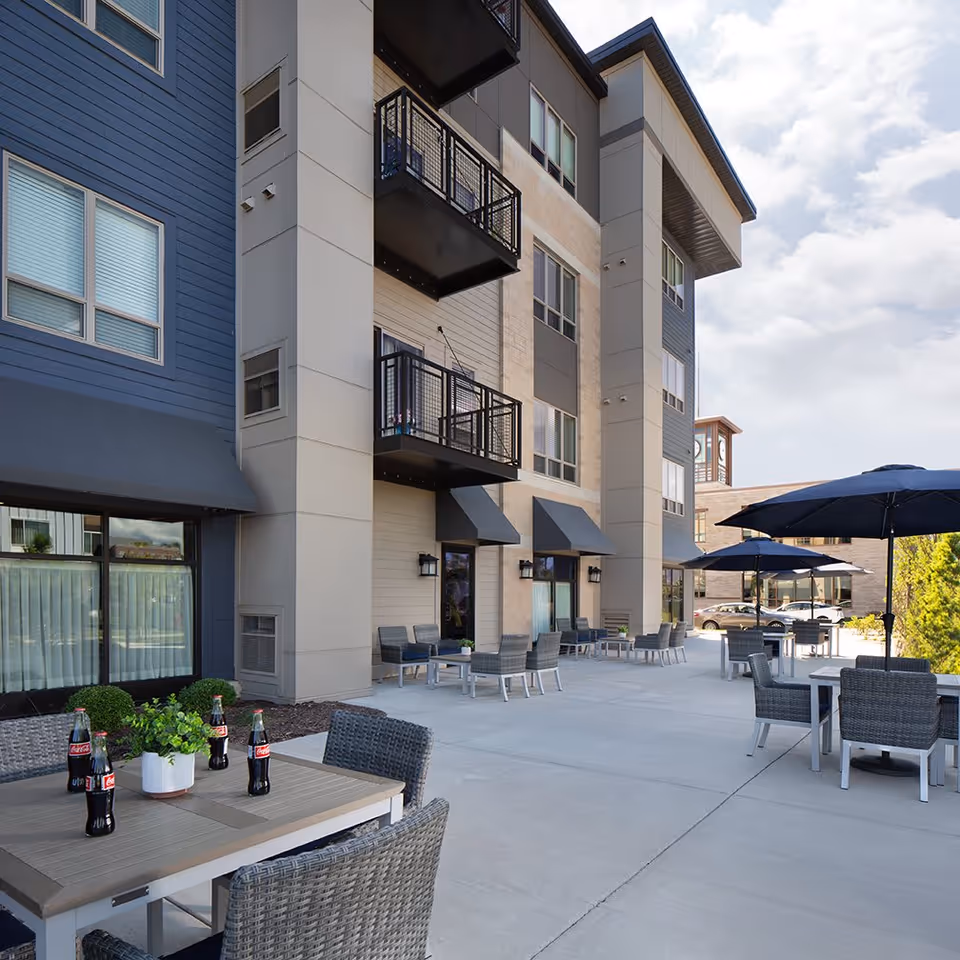 Outdoor patio area at The Waters of Oak Creek featuring multiple tables with wicker chairs and large blue umbrellas. The patio is adjacent to a multi-story building with balconies and awnings over the windows and doors. Several bottles of Coca-Cola and a small potted plant are on one of the tables.
