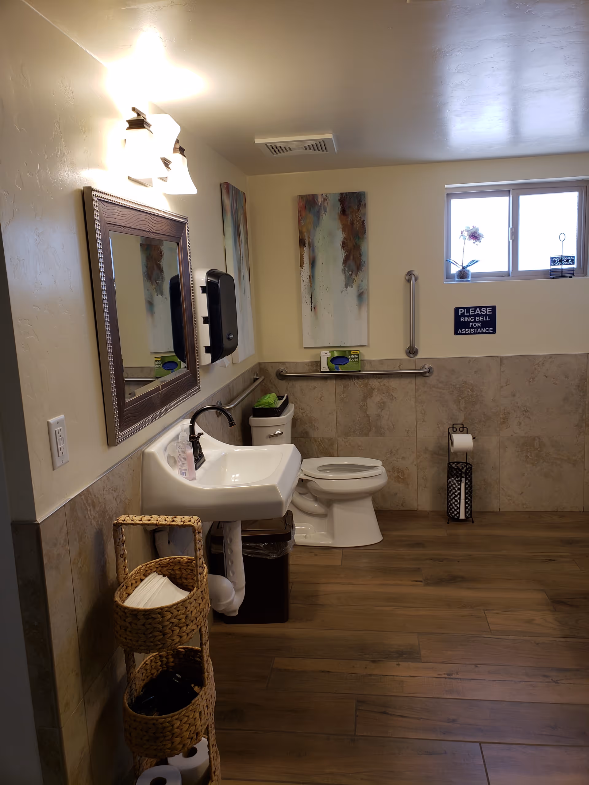 A clean and accessible bathroom with a white sink, a mirror above it, a toilet with grab bars on the wall, a small window with a flower pot, and a sign that reads 'PLEASE RING BELL FOR ASSISTANCE'. There is a wicker basket holding toilet paper and other supplies, and the floor is wooden with beige tiled walls.