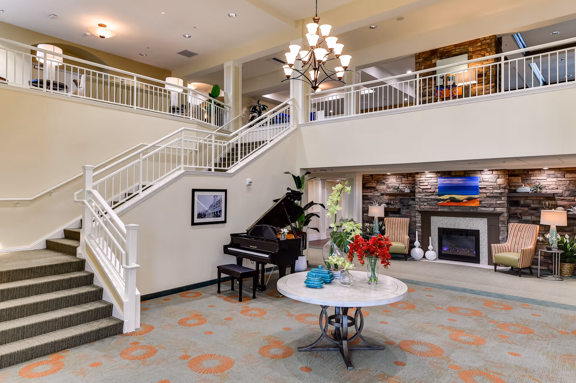 Spacious and well-lit senior living facility common area with a staircase leading to an upper level. The room features a grand piano, a round table with floral arrangements, a fireplace with stone surround, and comfortable armchairs. A chandelier hangs from the ceiling, and the carpet has a circular pattern in orange and beige tones.