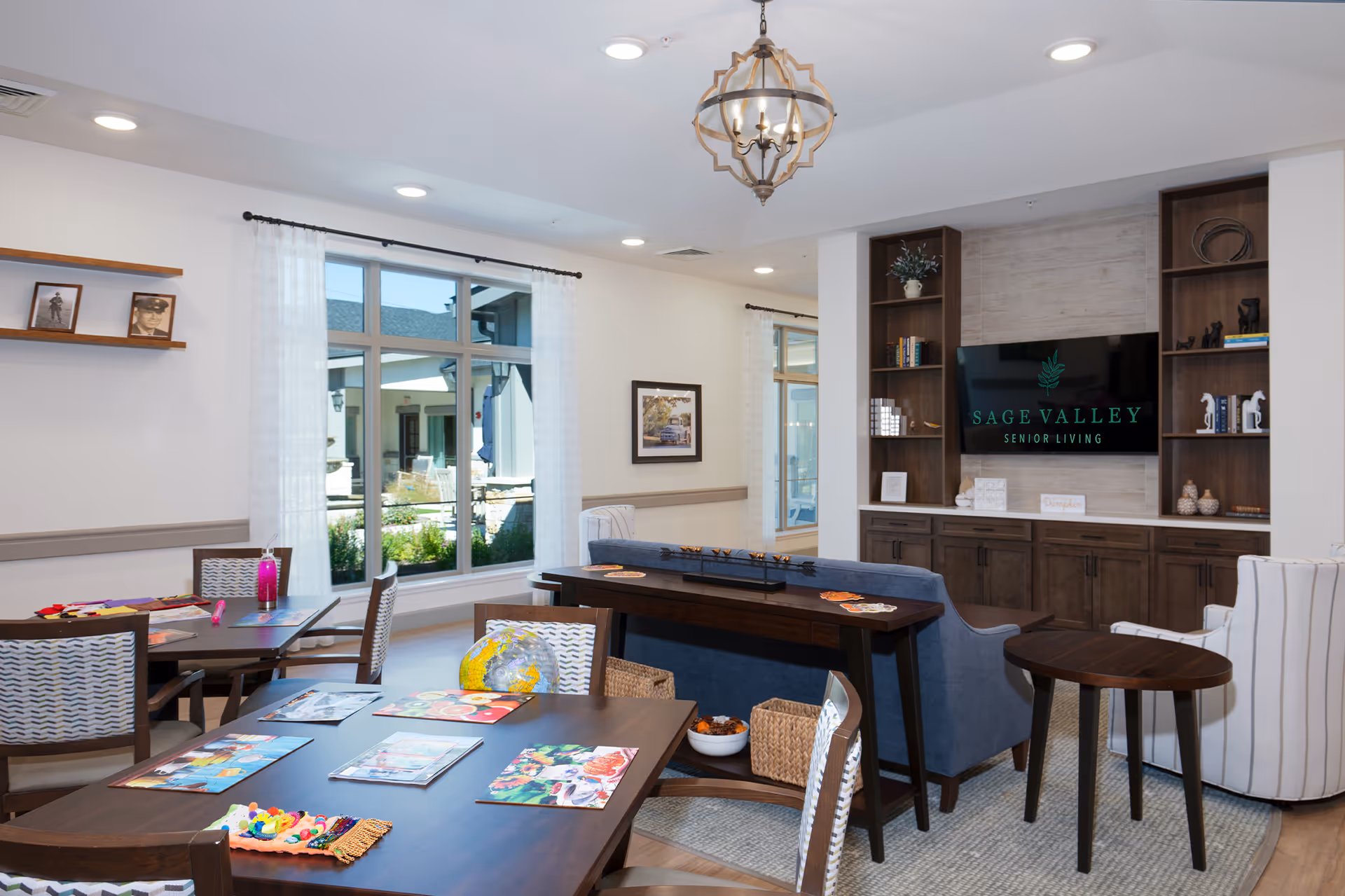 Well-lit senior living common room with activity tables, seating, and a TV displaying 'Sage Valley Senior Living'.