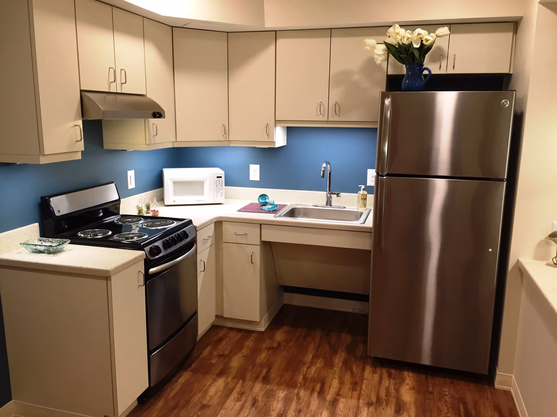 A compact kitchen with beige cabinets, a stainless steel refrigerator, an electric stove with four burners, a microwave on the countertop, a sink with a faucet, and a blue wall backsplash. There is a vase with white flowers on top of the refrigerator and a wooden floor.