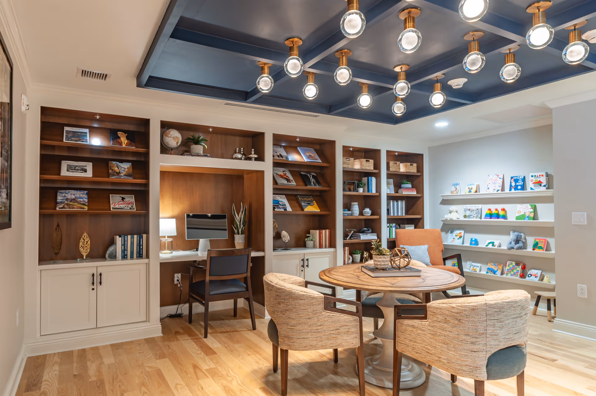 A cozy interior room with wooden bookshelves filled with books and decorative items, a small desk with a computer and chair, a round wooden table surrounded by four upholstered chairs, and a wall-mounted shelf displaying children's books and toys. The ceiling features a blue coffered design with multiple round light fixtures.