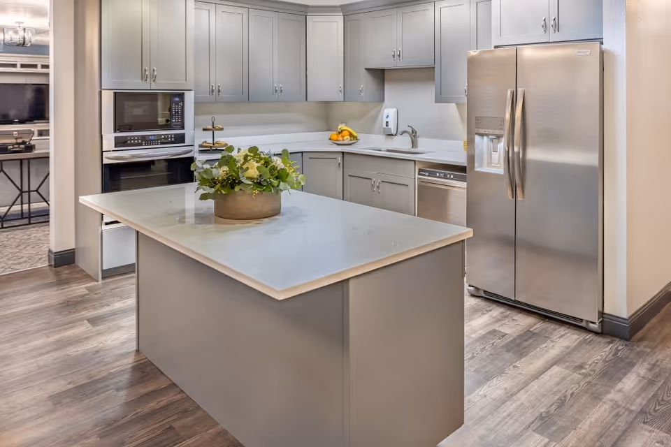Modern kitchen with gray cabinets, a large island with a flower arrangement, stainless steel refrigerator, built-in microwave and oven, dishwasher, and a bowl of fruit on the counter.
