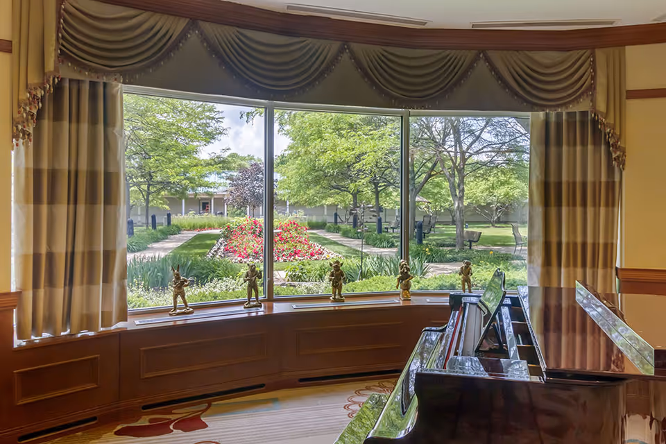 View from inside a room with a large bay window framed by striped curtains and a valance, overlooking a garden with green trees, a flower bed with red and white flowers, and walking paths. A polished grand piano is positioned near the window, with small decorative figurines placed on the window ledge.
