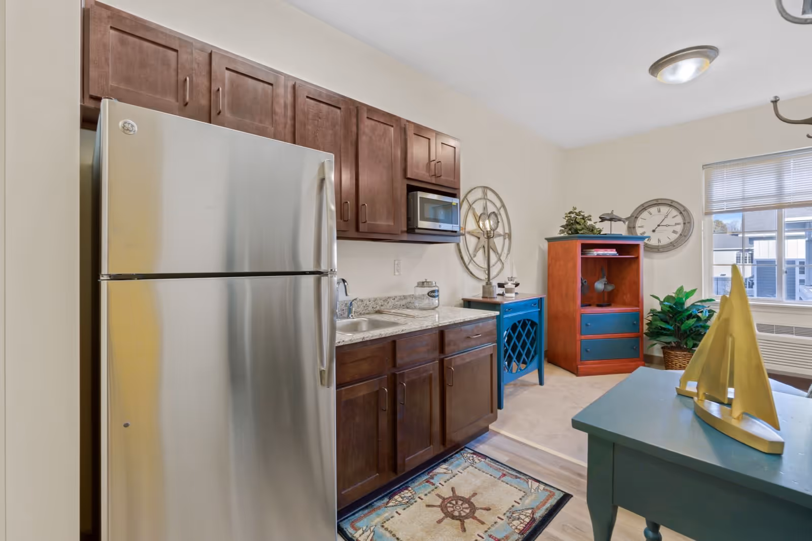 A small kitchen area with stainless steel refrigerator, dark wood cabinets, a microwave, and a sink. Adjacent to the kitchen is a living space with a blue table featuring a yellow sailboat decoration, a red and blue cabinet, a large wall clock, a potted plant, and a window with blinds letting in natural light.