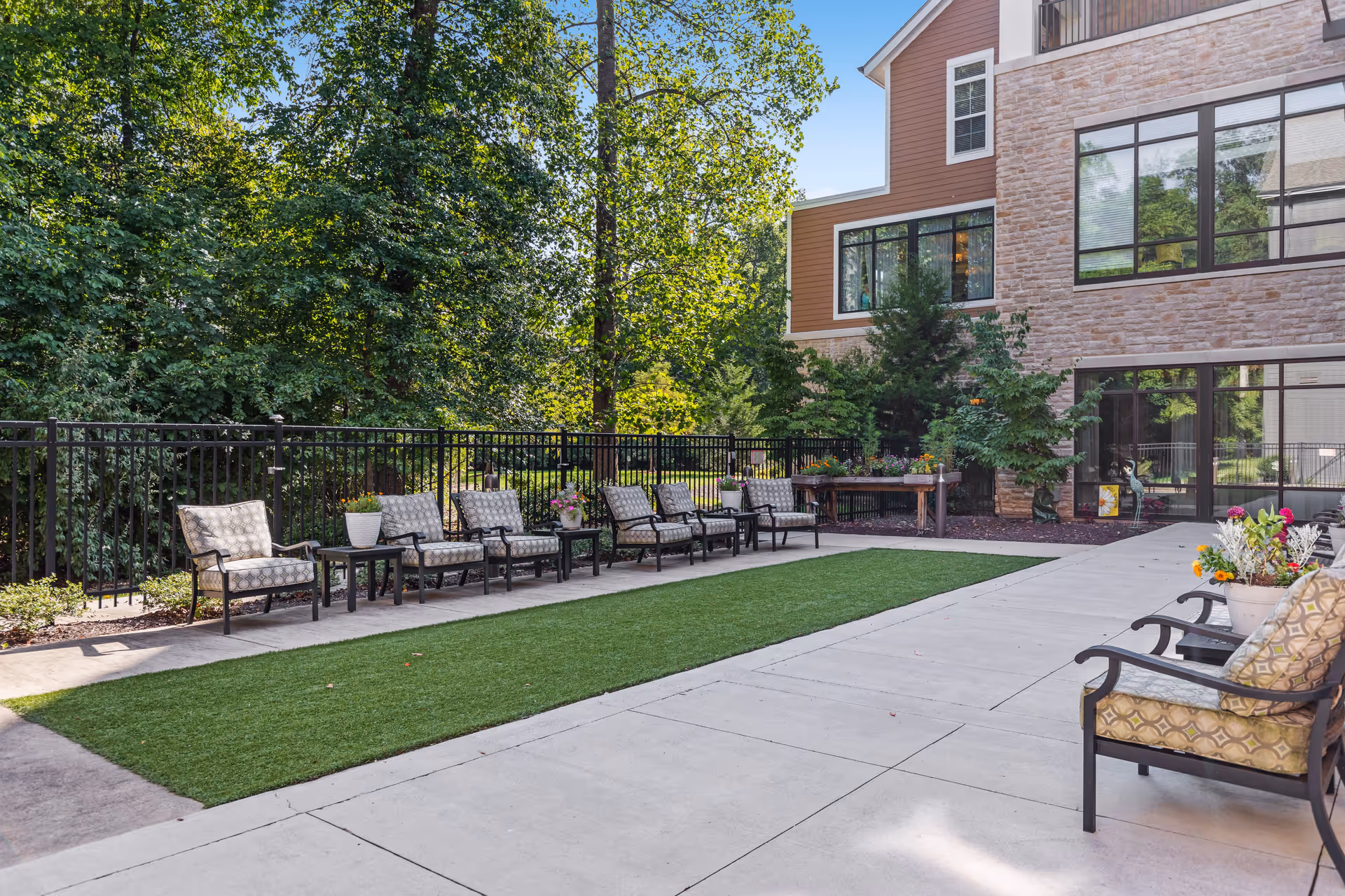 Outdoor patio area at Heritage Village Assisted Living and Memory Care featuring a row of cushioned chairs with small tables in between, a patch of artificial grass, and surrounding trees and greenery next to a multi-story building with large windows.